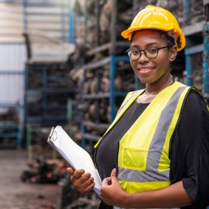 Une ouvrière afro-américaine vêtu d'un dossard jaune et d'un casque, debout dans une usine avec le sourire.