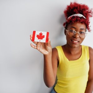 Femme afro-américaine aux cheveux afro, portant un chandail jaune et des lunettes, tenant le drapeau du Canada dans sa main.